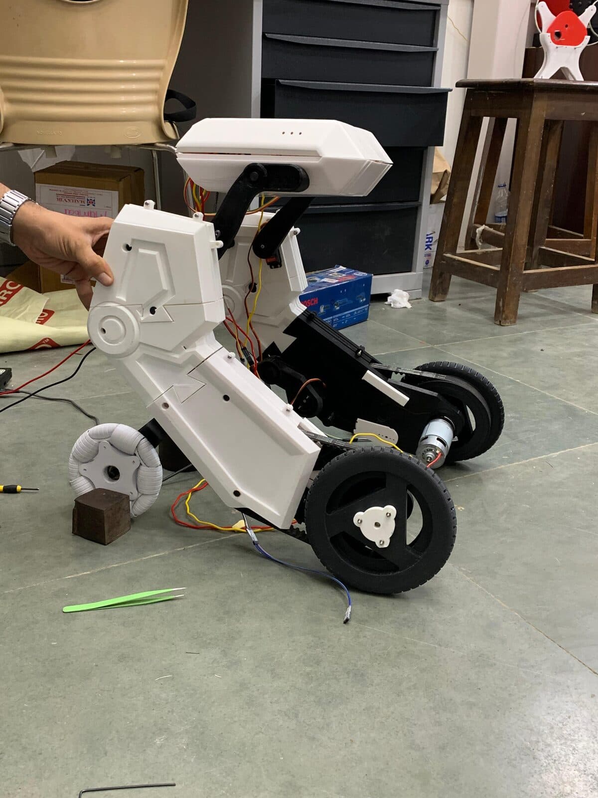 A half-scale assistive-mobility robot part-way through assembly on a workshop floor, its white shell open to show drive motors, belts and wiring.