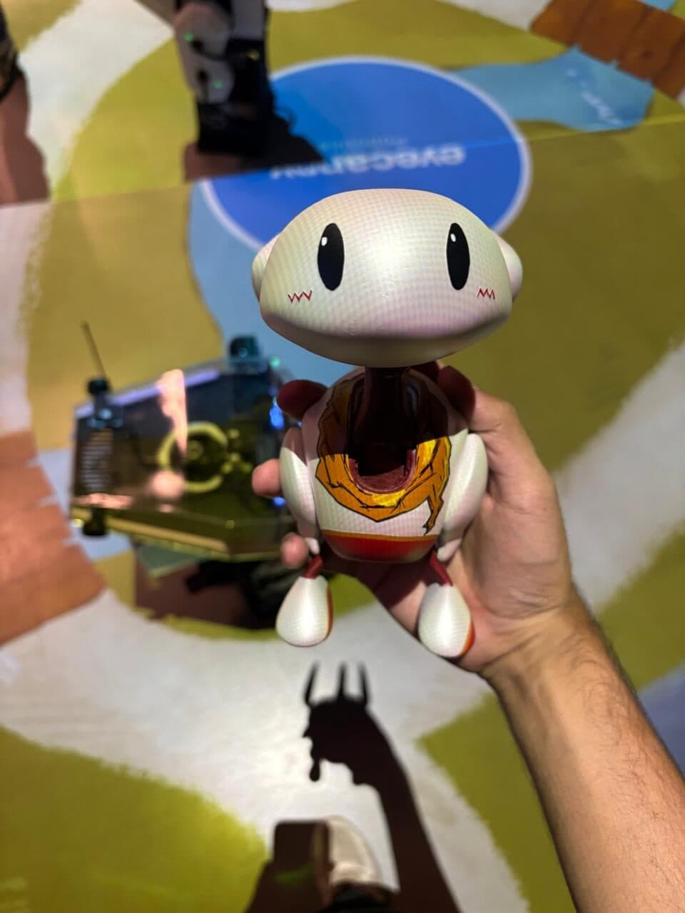 A small painted robot held in one hand above a printed floor graphic, with other robots out of focus behind it.