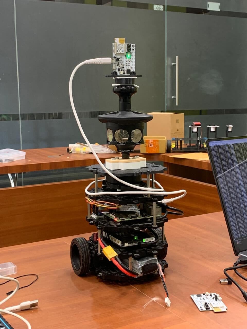 A small wheeled robot on a table carrying a stacked payload: compute boards on standoffs, a multi-lens sensor housing and a module on top.