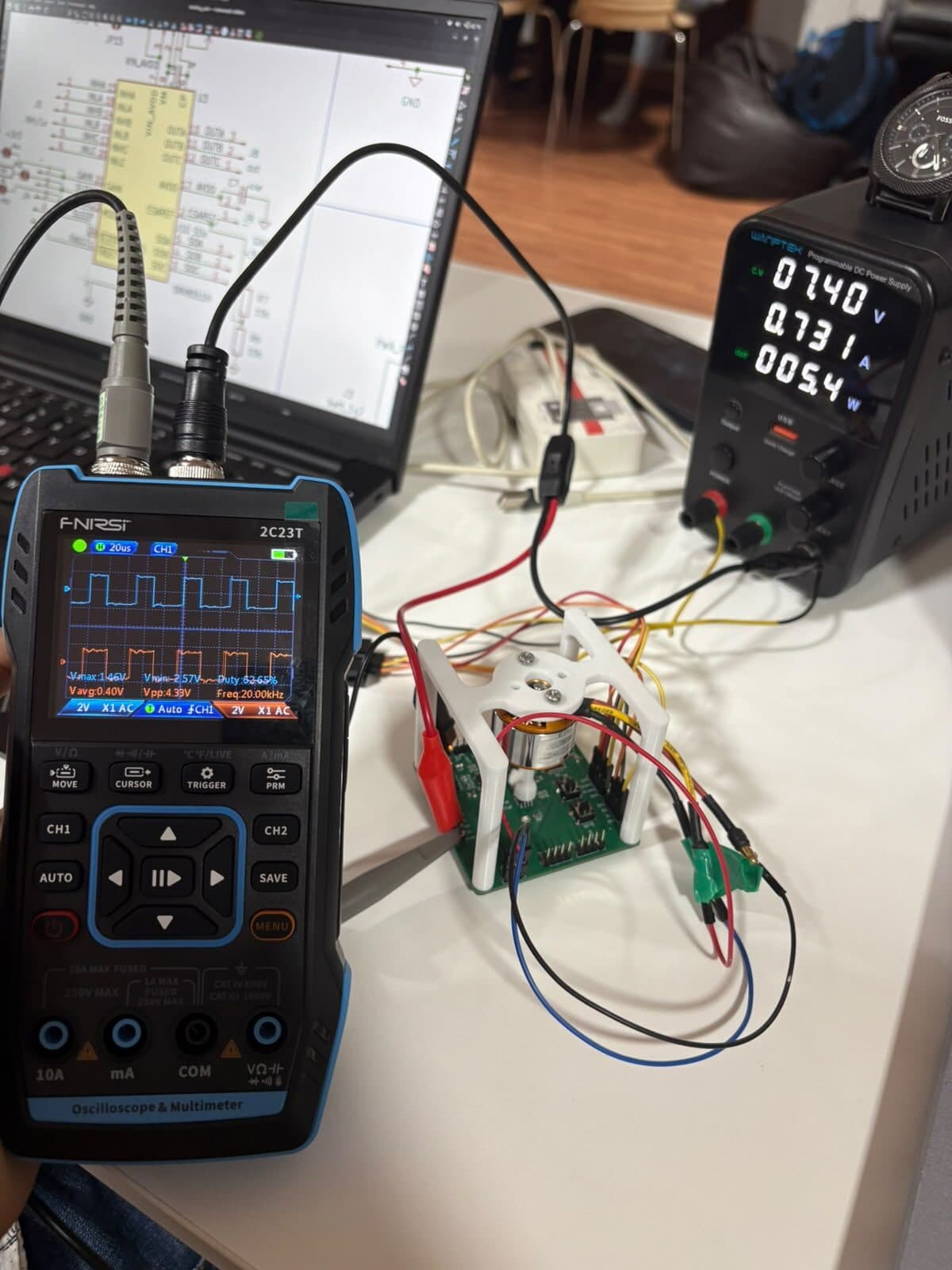 A handheld oscilloscope showing a 20 kHz PWM waveform, connected to a brushless motor on a driver board, with a bench power supply reading 7.40 volts and a schematic open on a laptop behind.