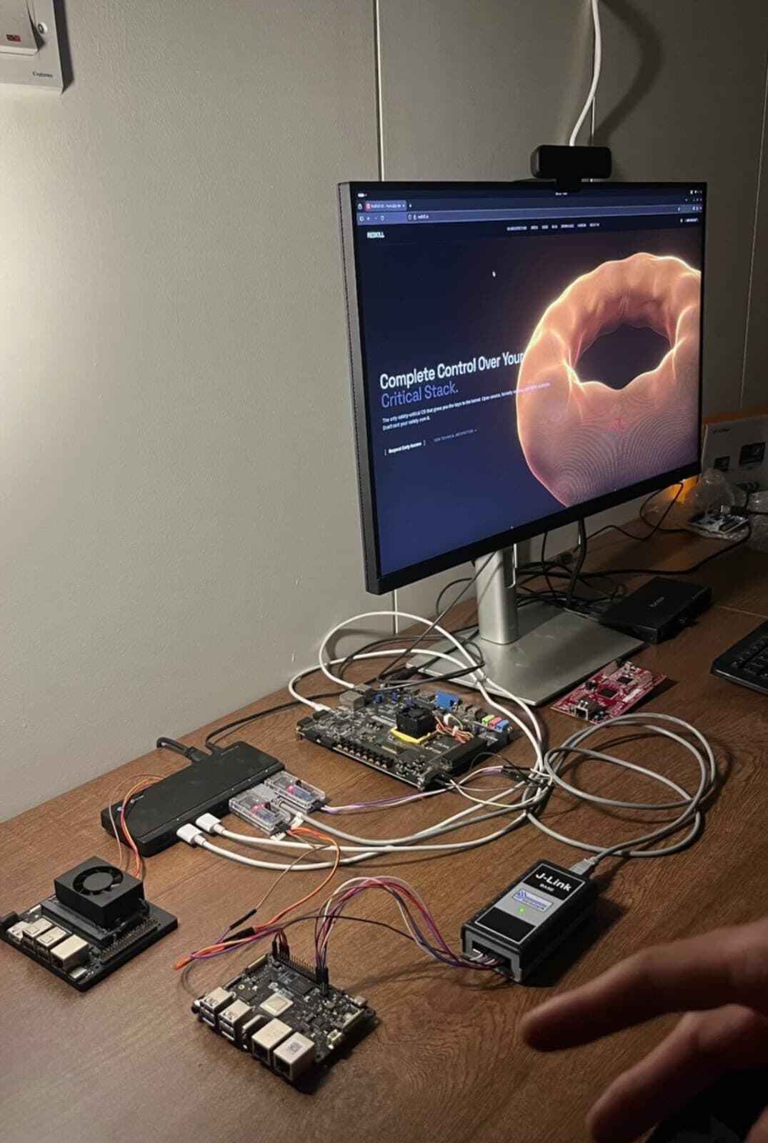 A desk with two single-board computers, a development board and a J-Link debug probe wired together, in front of a monitor showing the RedKill OS website.
